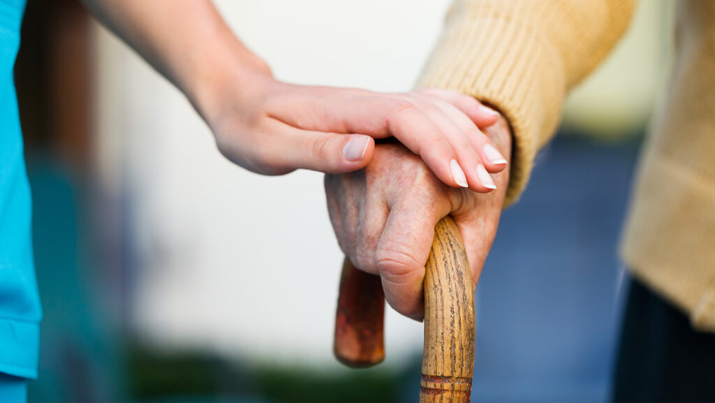 An older persons hand holding a walking stick with a younger personas hand on top.