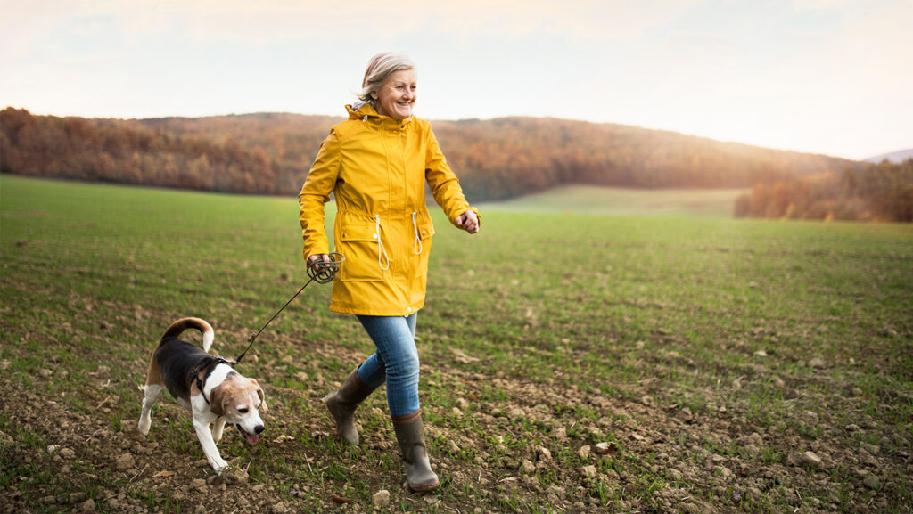 A lady in a bright yellow jacket walking her dog in a green field.